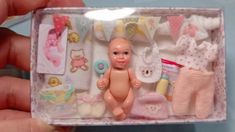 a hand holding a small doll in a box with clothes and baby items inside it