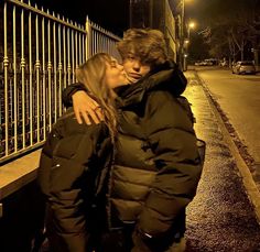 a man and woman standing next to each other on a sidewalk at night with their arms around each other