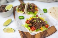 two tacos with meat, tomatoes and lettuce on a cutting board next to limes