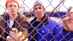 two men behind a chain link fence with one pointing at the camera and the other holding something in his hand