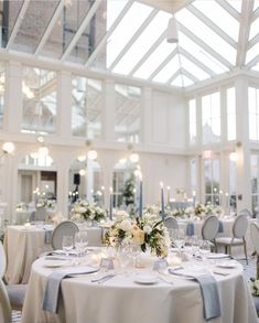 White round tables with hints of blue in the napkins and candles and a floral centerpiece in an atrium setting. Blue And White Flower Table Arrangements, Blue And White Round Table Decor, Wedding Table Decor Blue And White, Wedding Settings Indoor, Blue Round Table Wedding, Blue And White Table Setting Wedding, Dusty Blue Wedding Venue Decor, Blue And White Wedding Round Table Decor, Blue And White Wedding Reception Decor