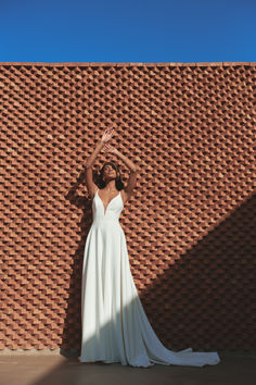 a woman standing in front of a brick wall wearing a white dress and holding her hands up