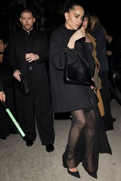 a woman in sheer pants and black jacket walking with her hand on her hip while another man stands behind her