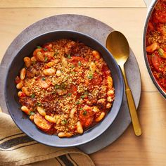 two pans filled with pasta and sauce on top of a wooden table next to a spoon