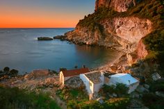 a small house sitting on top of a rocky cliff next to the ocean at sunset