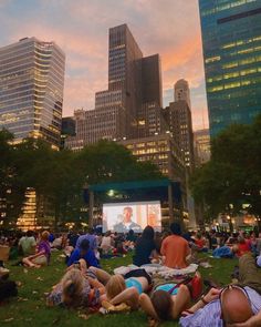 many people are sitting in the grass watching a movie on a big screen at dusk