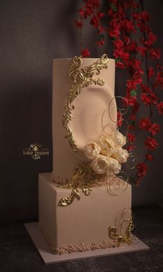 a three tiered white wedding cake with gold decorations and flowers on top, sitting in front of a red flower bush