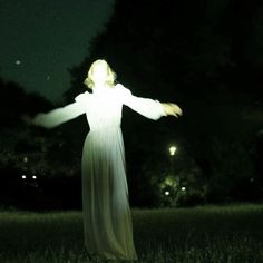 a woman standing in the grass with her arms spread out and lights on behind her