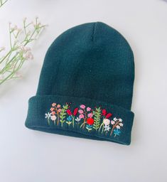 a green beanie with embroidered flowers on it next to some white and pink flowers