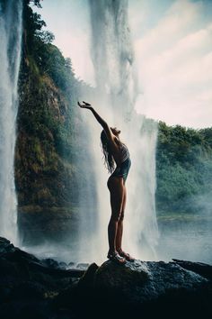 a woman standing on top of a rock in front of a waterfall with her arms outstretched