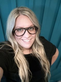 a woman with long blonde hair and black glasses posing for a photo in front of a blue curtain