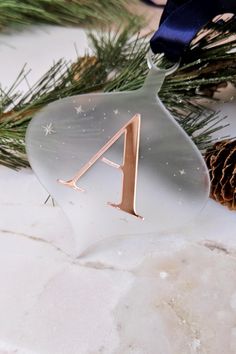 a glass ornament with the letter a on it next to pine cones and a blue ribbon