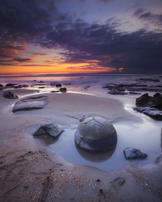 a large rock sitting on top of a sandy beach under a cloudy blue sky at sunset