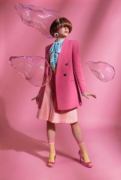 a woman in a pink coat and dress with wings on her head, standing against a pink background