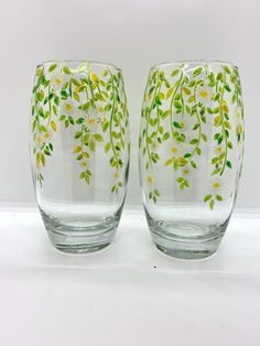 two glass vases sitting side by side on a white surface with green leaves and yellow flowers painted on them