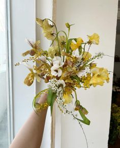 a hand holding a bouquet of flowers in front of a window with white and yellow flowers