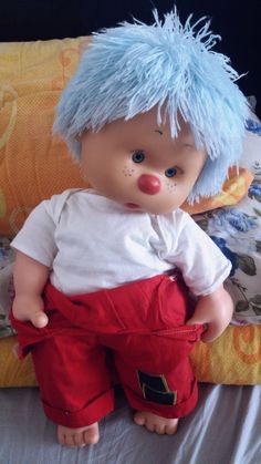 a small doll with blue hair and red pants on top of a bed next to pillows