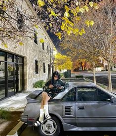 a woman sitting on top of a car parked in front of a building next to a tree