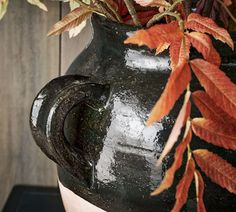 a black vase with red and green leaves in it sitting on a table next to a wooden wall