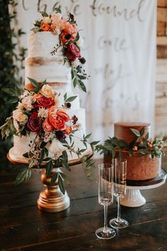 a wedding cake with flowers and greenery on top sits next to two champagne glasses