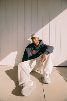 a man sitting on the ground wearing white pants and a blue hoodie, leaning against a wall