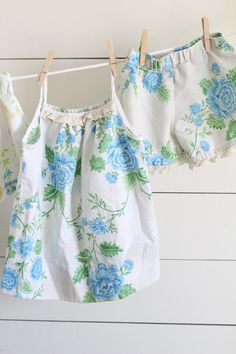 two baby blouses hanging on clothesline against white plank wall with blue flowers and green leaves