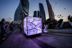 people are standing around in front of a purple cube on the ground with buildings in the background