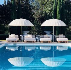 two lounge chairs with umbrellas next to a swimming pool in front of some trees