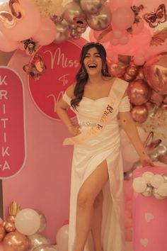 a woman in a white dress standing next to a pink wall with balloons and decorations