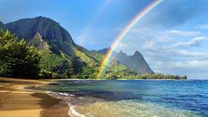 a rainbow shines in the sky over a beach with mountains and trees on either side