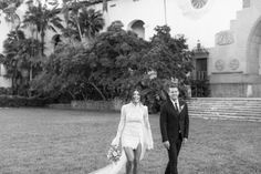 a bride and groom walk through the grass in front of a building with palm trees