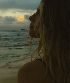 a woman standing on top of a sandy beach next to the ocean at sunset with her hair blowing in the wind