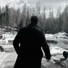 a man in a black coat is looking out at the snow covered ground and trees