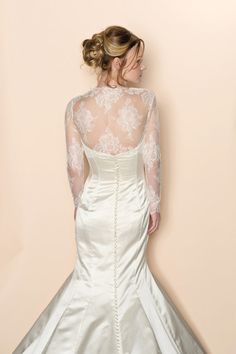 a woman in a white wedding dress with long sleeves and lace on her back, standing against a wall