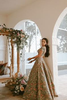 a woman in a floral dress leaning against a wall with flowers on the floor and an archway behind her