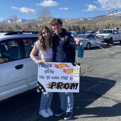 a man and woman standing in front of a white car holding a sign that says you're hot