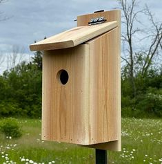 a wooden bird house on top of a green pole in the grass and flowers behind it