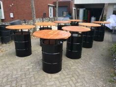 several round wooden tables sitting on top of a brick floor next to black barrels in front of a building