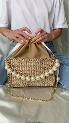 a woman sitting on the ground holding a straw bag with pearls hanging from it's sides