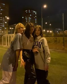 three girls posing for the camera in front of some tall buildings at night with their arms around each other