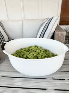 a white bowl filled with green beans sitting on top of a wooden table next to pillows