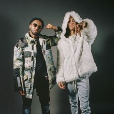 two people standing next to each other in front of a gray background wearing jackets and hoods