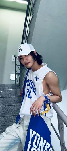 a young man wearing a white hat and tank top leaning against a stair rail with his hand on his hip