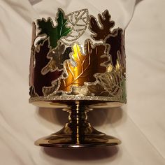 a gold and silver candle holder with leaves painted on the top, sitting on a white sheet