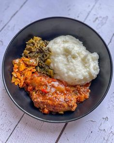 a black plate with mashed potatoes, meat and vegetables on it sitting on a white wooden table