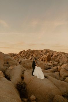 a bride and groom are standing on rocks in the desert at sunset, with their arms around each other