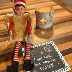 an elf sitting on top of a wooden table next to a cup and sign that says i got cold, but now'm toasty