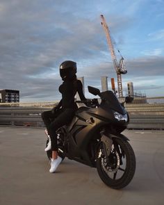 a woman sitting on top of a black motorcycle in front of a construction area with cranes