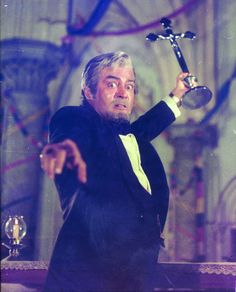 a man in a tuxedo holding a cross and pointing at something with his right hand