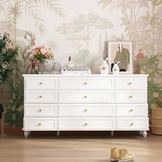 a white dresser sitting in front of a wall with flowers and plants on top of it